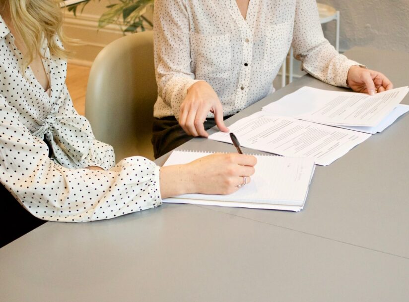 woman signing on white printer paper beside woman about to touch the documents